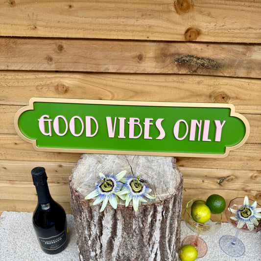 A green art deco inspired sign with the text “good vibes only” in light pink with a gold frame. Styled on a tiled work surface against a wooden wall with a bottle of sparkling wine, decorative cocktail glasses, limes and passionflowers.